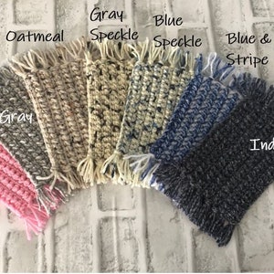 May include: A set of seven crocheted coasters in various colors and patterns, including pink, gray, oatmeal, gray speckle, blue speckle, blue and white stripe, and indigo. The coasters have fringe around the edges.