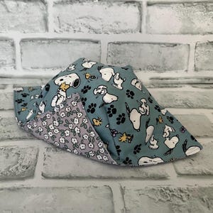 May include: A teal fabric bowl cosy with a Snoopy and paw print pattern on the outside and a floral pattern on the inside. The cosy is folded and ready to use. The background is a white brick wall.