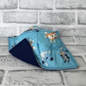 May include: A square fabric bowl cosy featuring a light blue exterior with a repeating pattern of cartoon dogs in various poses. The interior lining is a solid dark navy blue fabric. The item is constructed with stitched seams.