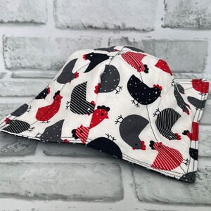 May include: A white fabric pot holder with a black and red chicken print. The pot holder is shaped like a triangle with a pointed top and a wide base.