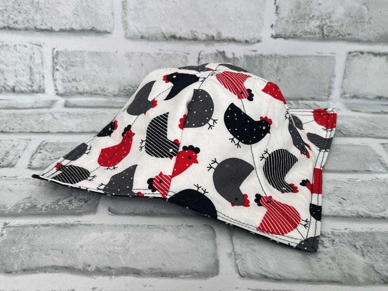 May include: A white fabric pot holder with a black and red chicken print. The pot holder is shaped like a triangle with a pointed top and a wide base.