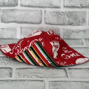 May include: Red and white Coca-Cola themed bowl cozy with a striped interior. The exterior features Coca-Cola logos and text. The interior has vertical stripes in red, black, green, and yellow. A decorative and functional kitchen accessory.
