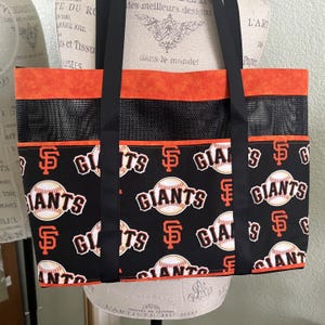 May include: A black tote bag featuring an orange top band, a black mesh middle section, and a black lower panel printed with the San Francisco Giants logo and baseball graphics. The bag includes two long black fabric shoulder straps.