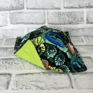 May include: A fabric bowl cosy featuring a black background with a colourful pattern of lizards, frogs, and tropical leaves. The interior of the quilted bowl holder is a solid lime green fabric, and the exterior displays a vibrant jungle print.