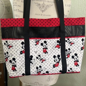 May include: A tote bag featuring a white fabric base with a repeating Mickey Mouse pattern, a black mesh middle section, and a red top border with black polka dots. The bag includes two black fabric shoulder straps and red trim accents.
