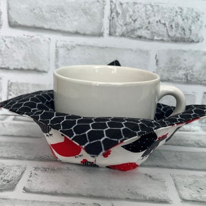 May include: A white ceramic mug sits inside a black and white fabric cup cosy with a chicken print. The cosy has a black and white checkered pattern on the inside and a white background with red and black chickens on the outside.