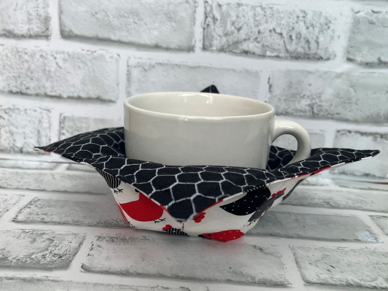 May include: A white ceramic mug sits inside a black and white fabric cup cosy with a chicken print. The cosy has a black and white checkered pattern on the inside and a white background with red and black chickens on the outside.