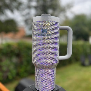 Puede incluir: Una taza de viaje gris con asa, cubierta de piedras de cristal iridiscentes.