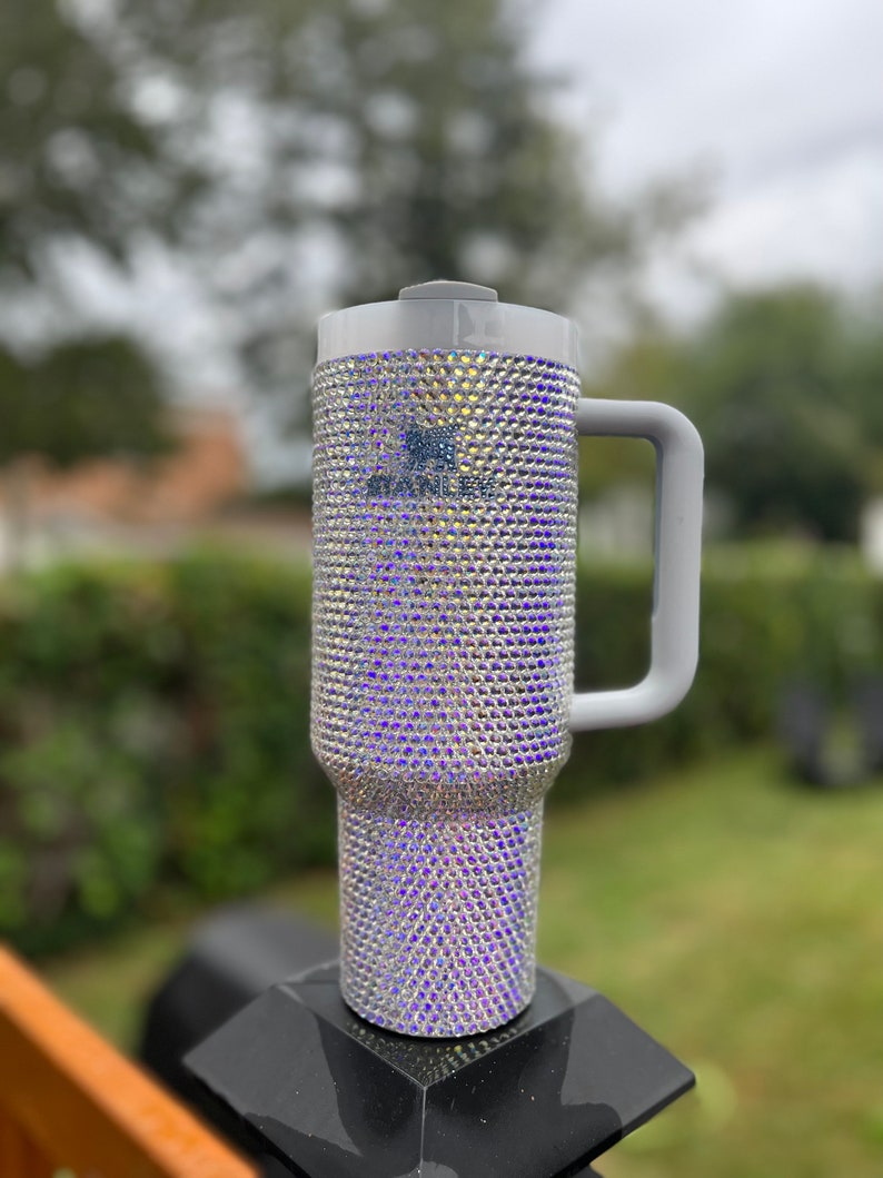 Puede incluir: Una taza de viaje gris con asa, cubierta de piedras de cristal iridiscentes.