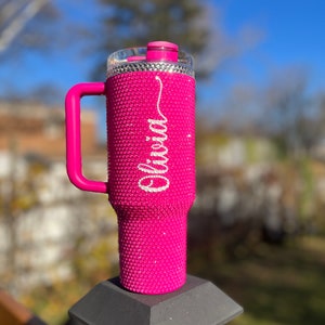 Puede incluir: Un vaso rosa cubierto de diamantes de imitaci&oacute;n con asa y tapa blanca. El vaso tiene el nombre "Olivia" escrito en diamantes de imitaci&oacute;n blancos.