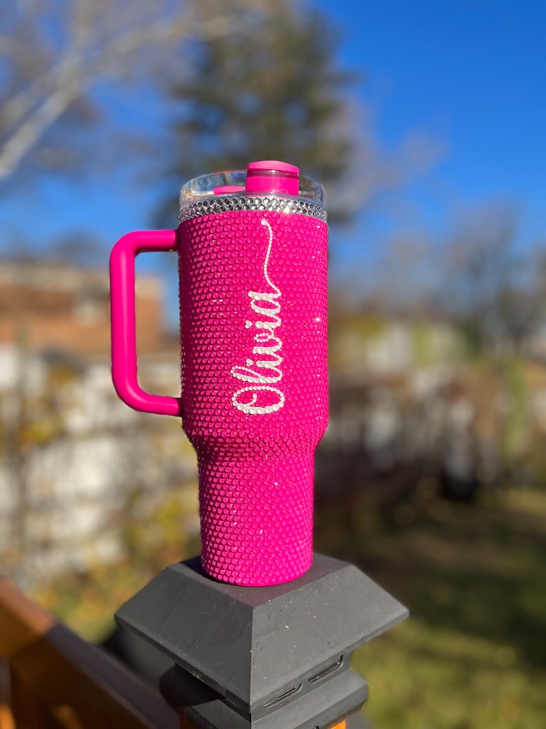 Puede incluir: Un vaso rosa cubierto de diamantes de imitaci&oacute;n con asa y tapa blanca. El vaso tiene el nombre "Olivia" escrito en diamantes de imitaci&oacute;n blancos.
