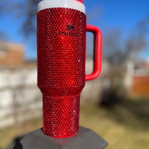 Puede incluir: Un vaso rojo cubierto de pedrer&iacute;a con asa roja y tapa blanca. El vaso est&aacute; sobre una superficie negra.