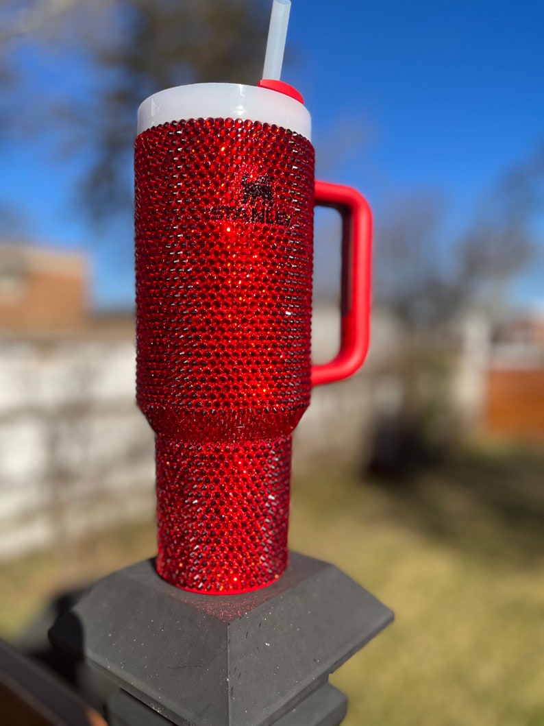 Puede incluir: Un vaso rojo cubierto de pedrer&iacute;a con asa roja y tapa blanca. El vaso est&aacute; sobre una superficie negra.