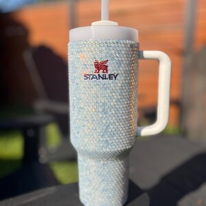 Puede incluir: Un vaso de acero inoxidable blanco con tapa transparente y una pajita. El vaso est&aacute; cubierto de piedras de cristal iridiscentes y tiene el logotipo de Stanley en la parte delantera.