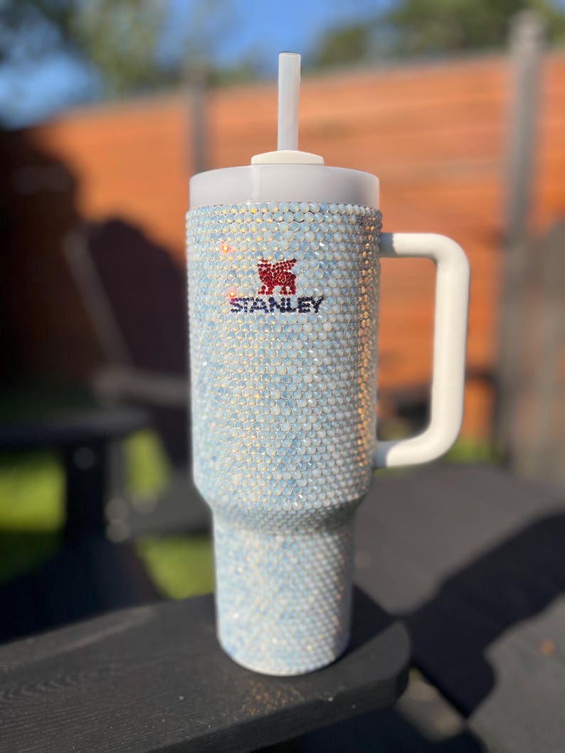 Puede incluir: Un vaso de acero inoxidable blanco con tapa transparente y una pajita. El vaso est&aacute; cubierto de piedras de cristal iridiscentes y tiene el logotipo de Stanley en la parte delantera.