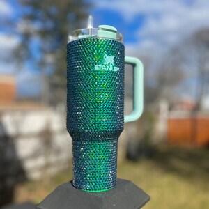 Puede incluir: Un vaso de acero inoxidable verde y azul con tapa y asa de color verde claro. El vaso est&aacute; cubierto de diamantes de imitaci&oacute;n y tiene la palabra "Stanley" impresa en el lateral.