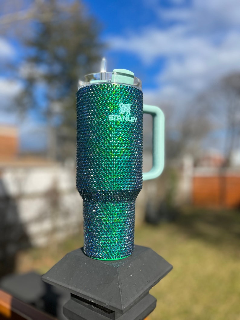 Puede incluir: Un vaso de acero inoxidable verde y azul con tapa y asa de color verde claro. El vaso est&aacute; cubierto de diamantes de imitaci&oacute;n y tiene la palabra "Stanley" impresa en el lateral.