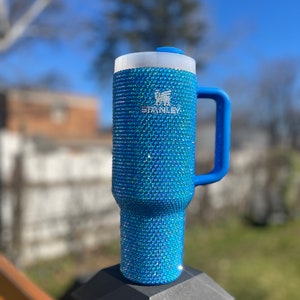 Puede incluir: Una taza de viaje Stanley azul con asa, cubierta de brillantes azules iridiscentes. La taza tiene un logotipo blanco que dice "Stanley" en el lateral.
