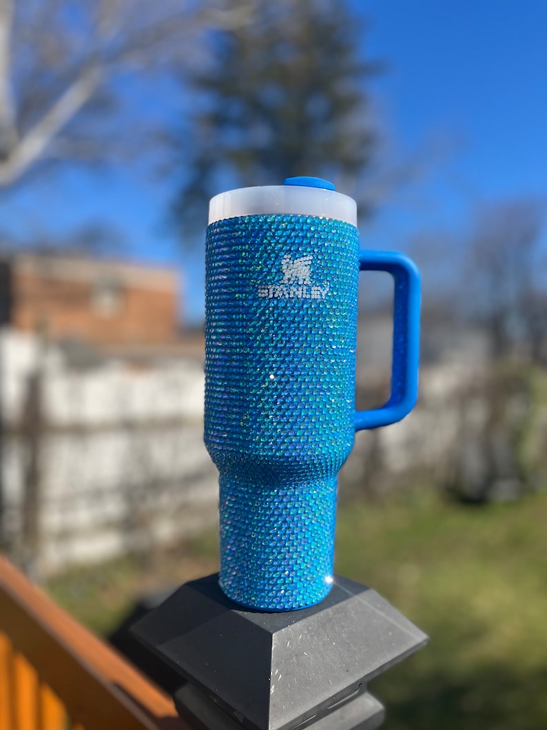 Puede incluir: Una taza de viaje Stanley azul con asa, cubierta de brillantes azules iridiscentes. La taza tiene un logotipo blanco que dice "Stanley" en el lateral.