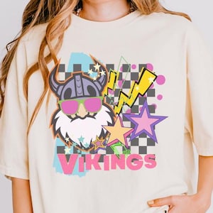 May include: A white t-shirt with a graphic design featuring a cartoon viking wearing sunglasses, a checkered pattern, lightning bolts, and stars. The text "VIKINGS" is printed in pink letters.