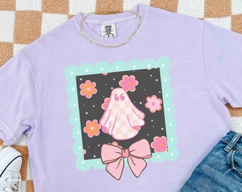 May include: A lilac t-shirt with a graphic of a ghost in a checkered pattern, surrounded by flowers and a pink bow. The ghost graphic is set within a square frame with a scalloped edge. A gold chain necklace is visible.