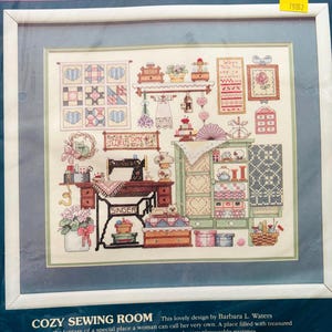 May include: A Sunset brand stamped cross stitch kit titled Cozy Sewing Room. The package features a printed design of a sewing room with a Singer sewing machine, a cabinet, quilts, and various sewing supplies. The kit includes cotton thread.