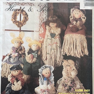 Puede incluir: Patrón de McCall's Crafts con varias muñecas de trapo hechas a mano, incluyendo una muñeca conejito y una muñeca novia. Las muñecas están hechas con hilo y tela, con detalles como sombreros y delantales. El patrón incluye el texto "Heart & Home."