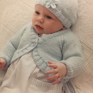 May include: A light blue knitted baby cardigan with a button closure and a matching light blue knitted hat with a white flower embellishment. The baby is wearing a white dress with a pocket.