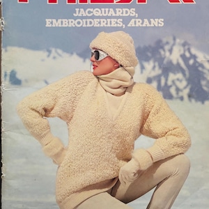 May include: A vintage magazine cover with the word "PHILDAR" in red, featuring a woman in cream-colored knitwear, including a sweater, hat, and trousers. The cover also includes the text "JACQUARDS, EMBROIDERIES, ARANS". The background shows a snowy mountain scene.