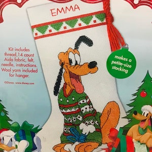 May include: Cross stitch stocking kit featuring a cartoon dog wearing a green and red sweater. The kit includes 14 count Aida fabric, felt, needle, instructions, and wool yarn. The stocking is 10 inches (25.4 cm) long and makes a petite-size stocking. The kit is titled "Emma".