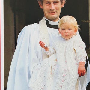 May include: A person in a white robe holds a baby wrapped in a white knitted blanket with pink floral accents. The baby is wearing a matching knitted outfit and holding a small silver object. The background is a blurred outdoor setting.