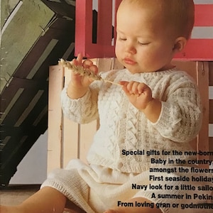 May include: A baby wearing a white knitted sweater and skirt sits on a wooden chair. The baby is holding a stalk of wheat in their hand. The image is from a magazine called "Pingouin" and the text "Spring-Summer" is visible in the top right corner.