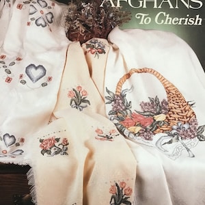 May include: Book 13 of the RainDrop Afghans To Cherish series. The book cover features a close-up of four crocheted afghans with floral designs. The afghans are white and cream with blue, pink, and green embroidery.