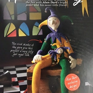 May include: A knitted jester doll wearing a purple and green costume, holding a smaller jester doll. The dolls are sitting on a wooden chest with a chequered floor in the background. The text "April Fool's Day" is at the top of the image. The text "Alan Dart exclusive design!" is in a circle at the bottom right of the image.