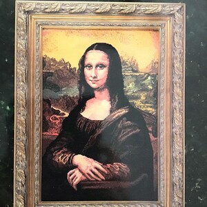 Könnte beinhalten: Ein Zählkreuzstich-Muster der Mona Lisa von Leonardo da Vinci. Das Muster ist in einem goldenen Rahmen gerahmt und hat einen grünen Hintergrund. Der Text "The Mona Lisa" ist in schwarzer Schrift oben auf dem Muster gedruckt. Der Text "by Leonardo da Vinci" ist in kleinerer schwarzer Schrift unter dem Titel gedruckt. Der Text "Counted Cross Stitch" ist in schwarzer Schrift unten links auf dem Muster gedruckt. Der Text "Adaptation by Rebecca Trent" ist in schwarzer Schrift unten rechts auf dem Muster gedruckt.