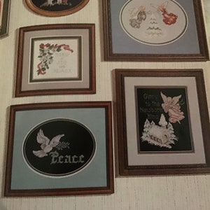 Puede incluir: Una colecci&oacute;n de dise&ntilde;os de bordado de punto de cruz enmarcados que se exhiben en una pared. Los dise&ntilde;os incluyen un cordero, &aacute;ngeles, una paloma con una rama de acebo y una escena de invierno con una iglesia. El texto en los marcos dice "Peace", "Glory to the Newborn King", y "Always in our Hearts and Remembered".