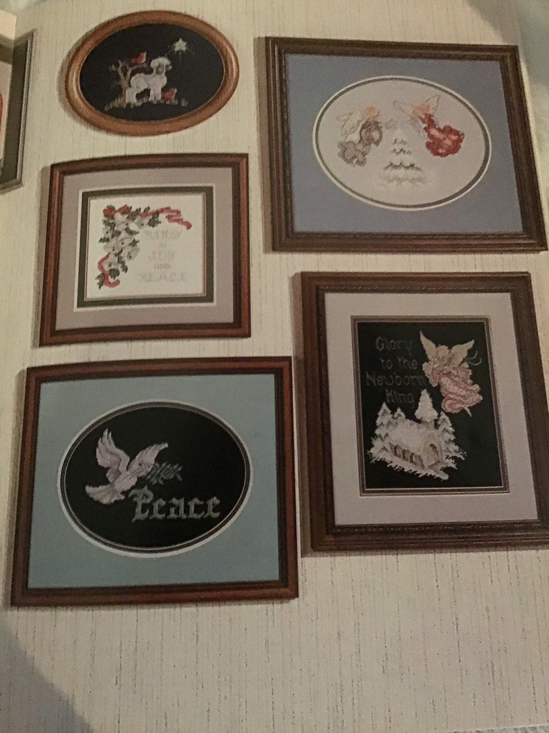 Puede incluir: Una colecci&oacute;n de dise&ntilde;os de bordado de punto de cruz enmarcados que se exhiben en una pared. Los dise&ntilde;os incluyen un cordero, &aacute;ngeles, una paloma con una rama de acebo y una escena de invierno con una iglesia. El texto en los marcos dice "Peace", "Glory to the Newborn King", y "Always in our Hearts and Remembered".