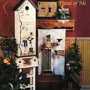 Puede incluir: Una colección de artesanías de madera pintadas con un tema campestre. Las artesanías incluyen una casita para pájaros, un estante, una figurita, un letrero y una casita pequeña. Las artesanías están pintadas en varios colores, incluyendo rojo, blanco, azul y verde. Las artesanías están dispuestas en un estante de madera y una alfombra. El título del libro es "Patches & Pieces of Me" de Nona Gabel.