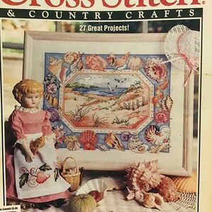 Könnte beinhalten: Ein Magazin-Cover für Cross Stitch & Country Crafts mit einem Kreuzstich-Design eines Muschelrandes mit einer Strandszene im Inneren. Das Magazin-Cover zeigt auch eine Porzellanpuppe, die ein rosa Kleid und eine weiße Schürze mit einem Kreuzstich-Design von Früchten trägt.