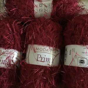 Pode incluir: Três novelos de fio vermelho-bordo com textura felpuda. O fio é etiquetado como "Moda Dea Prima" e tem uma etiqueta com o número de cor 1595, o peso do fio e o tamanho das agulhas.
