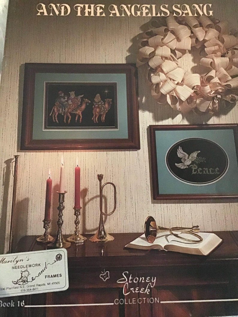 Puede incluir: Un libro titulado "And The Angels Sang" con una etiqueta de Stoney Creek Collection. El libro est&aacute; abierto y encima hay una trompeta dorada. Hay tres candelabros con velas rojas y rosas sobre una mesa de madera. Una imagen de punto de cruz enmarcada de los tres reyes magos montando camellos est&aacute; en la pared. Una imagen de punto de cruz enmarcada de una paloma con la palabra "Paz" est&aacute; en la pared. Una corona hecha de papel blanco est&aacute; en la pared.