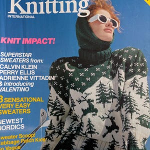 Könnte beinhalten: Ein Magazincover für Vogue Knitting International, das eine Frau zeigt, die einen grün-weißen Strickpullover mit einer Winterwald-Szene trägt. Der Titeltext lautet "Knit Impact!" und "Superstar Sweaters from: Calvin Klein, Perry Ellis, Adrienne Vittadini & Introducing Valentino".