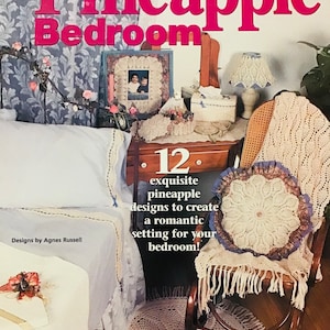 Op de afbeelding: Een haakpatroonboek met de titel "Pineapple Bedroom" met 12 prachtige ananaspatronen om een romantische sfeer in uw slaapkamer te creëren. De omslag toont een gehaakte deken, een kussen en een lampenkap in een slaapkamersetting.
