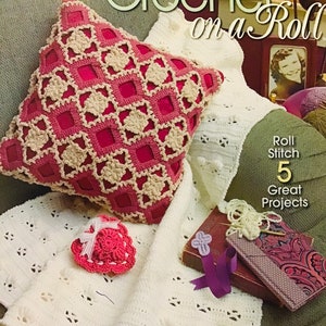 May include: A crochet book titled "How to Crochet on a Roll" with a price tag of £6.99. The book cover features a white crocheted blanket with a pink crocheted pillow and a pink crocheted heart-shaped object.