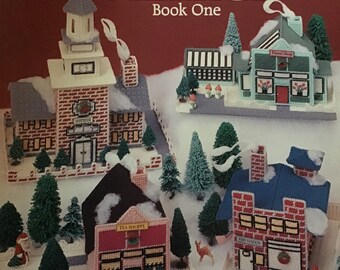 May include: Plastic canvas pattern for a Christmas village, featuring a red brick fire station, a pink tea shop, a green floral shop, and a white church. The pattern is titled "Home for Christmas Village" and is by Don FranzMeier.