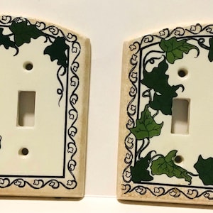 May include: Two white ceramic light switch covers with a green ivy design. The covers have a black border and a decorative scroll design.