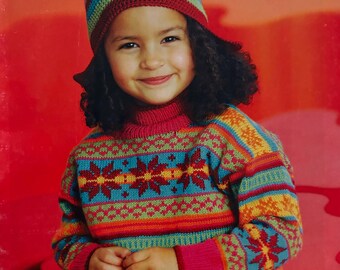 May include: A young girl wearing a colourful knitted sweater with a snowflake pattern and a multi-coloured crocheted hat. The sweater has a pink turtleneck and the hat has a red brim. The girl is smiling and has curly brown hair. The background is a solid red.