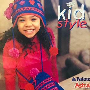 May include: A young girl wearing a blue and red knitted hat with hearts and pom-poms. She is also wearing a pink sweater and a blue and red knitted bag with pom-poms. The image is from a knitting pattern book called "Patons Kid Style".