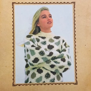 Könnte beinhalten: Ein weißer Pullover mit einem grünen und braunen Blatt-Muster. Der Pullover ist aus Wolle und hat einen hohen Kragen. Der Text "Álafoss Lopi" ist unter dem Bild gedruckt.