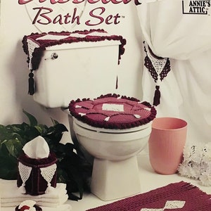 May include: Crochet pattern for a bathroom set featuring a toilet seat cover, toilet tank cover, tissue box cover, and bath mat. The set is crafted in a burgundy colour with white accents and tassels.